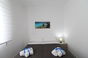Een hotelkamer met een bed, twee handdoeken vastgebonden met blauwe linten, een nachtkastje met een lamp en een lijst met een kustlandschap.
