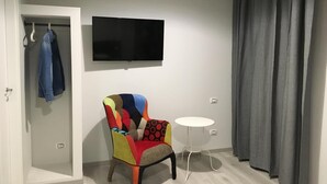 Una habitación con un sillón colorido, una pequeña mesa blanca, un televisor montado en la pared y una puerta con un abrigo colgado.
