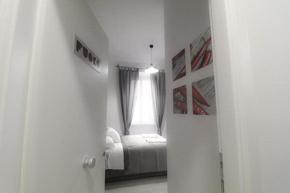 En smal hall leder in till ett sovrum med en säng, ett fönster med gardiner och tavlor med bilder av chilipeppar som hänger på väggen.