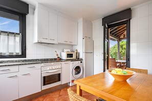 Una cocina moderna con armarios blancos, microondas, refrigerador y lavadora. Hay una mesa de madera con un bol de fruta.