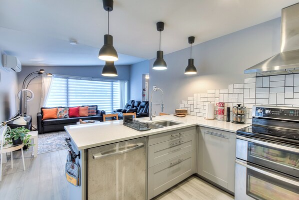 Une cuisine moderne dotée d’un îlot central, d’appareils électroménagers en acier inoxydable, de luminaires suspendus et offrant une vue sur un espace de vie avec un canapé et des fauteuils.