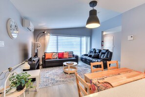 Un salon moderne avec un canapé, une table à manger, une lampe et une grande fenêtre équipée de stores.
