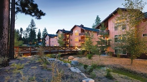 Un hôtel à plusieurs étages doté de balcons, entouré d’arbres et surplombant un pont en bois.