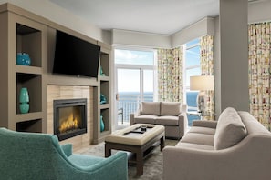 A modern living room with a fireplace, comfortable seating, a flat-screen TV, and a view of the ocean.