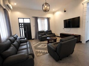 A living room with a sectional sofa, a recliner, a coffee table, a flat-screen TV, and a large window with curtains. - Your Hidden Escape Near Kilimanjaro 🌿 (Boma Ng'ombe)