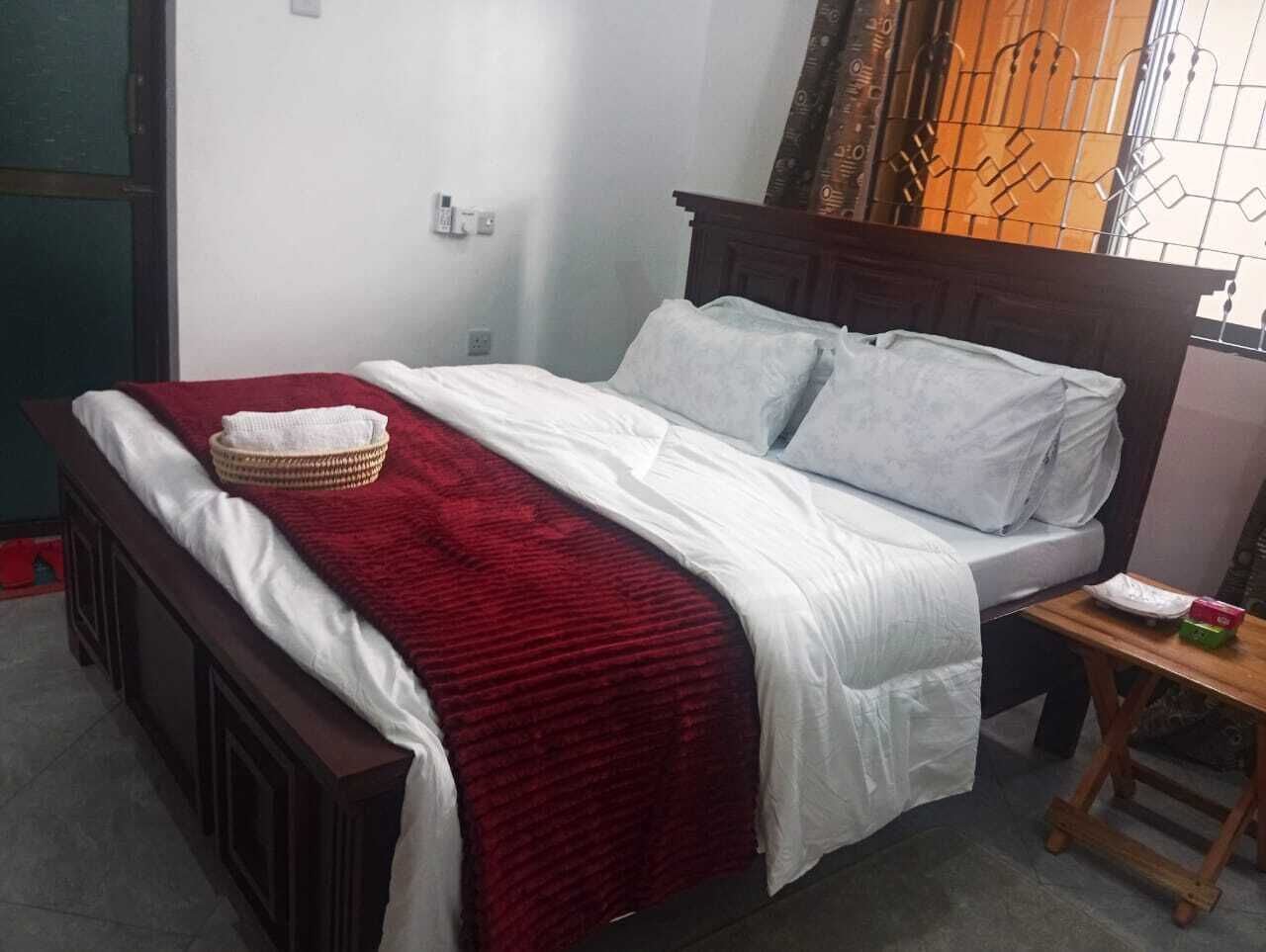Un lit simple avec une couverture rouge, des draps blancs et une tête de lit en bois. Une petite table en bois se trouve à côté du lit, et un objet blanc y est posé.