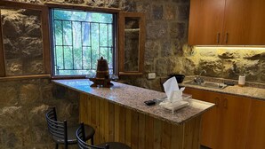 A kitchen with a stone wall, wooden cabinets, a granite countertop, a window with a view, and a bar area with stools.