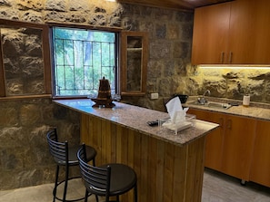 A kitchen with a stone wall, wooden cabinets, a granite countertop, a window with a view, and a bar area with stools.