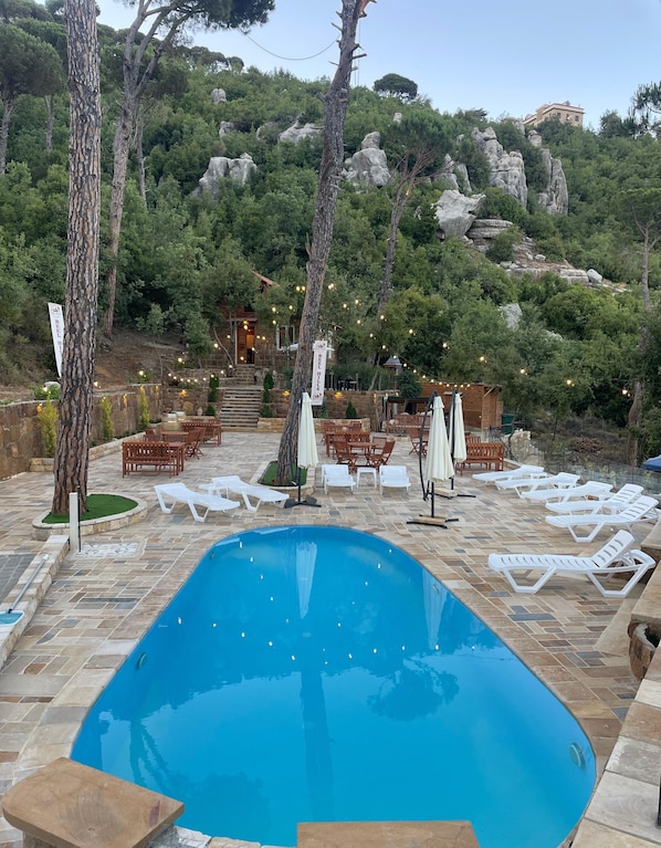 A swimming pool with lounge chairs and tables, surrounded by trees and a stone building.