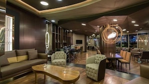 Um hall de hotel moderno com um sofá, poltronas, uma mesa de centro de madeira e uma área de refeições com cadeiras e uma mesa.