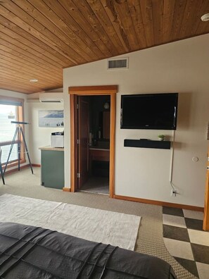 A room with a wooden ceiling, a bed, a television mounted on the wall, and a window with a view.