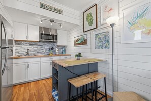 Eine Küche mit weißen Schränken, einer Mikrowelle und einer Arbeitsplatte mit Spüle. An der Wand befinden sich gerahmte Bilder und ein Schild mit der Aufschrift 'Beach House'.