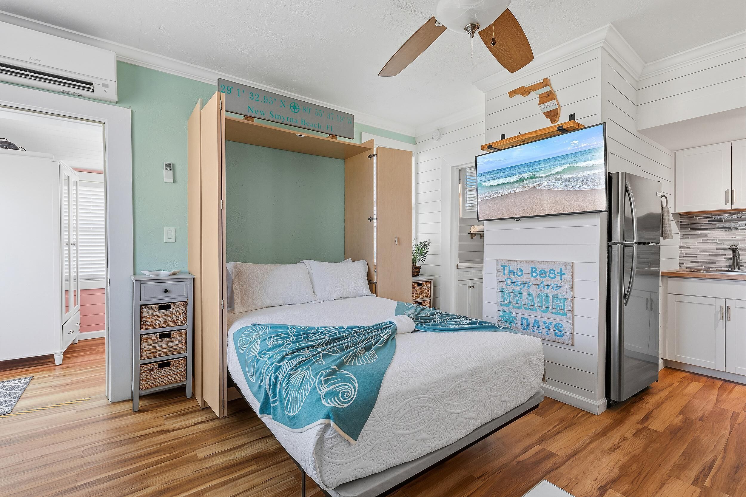 A bedroom with a bed, a ceiling fan, a wall-mounted TV, and a kitchen area with a refrigerator.