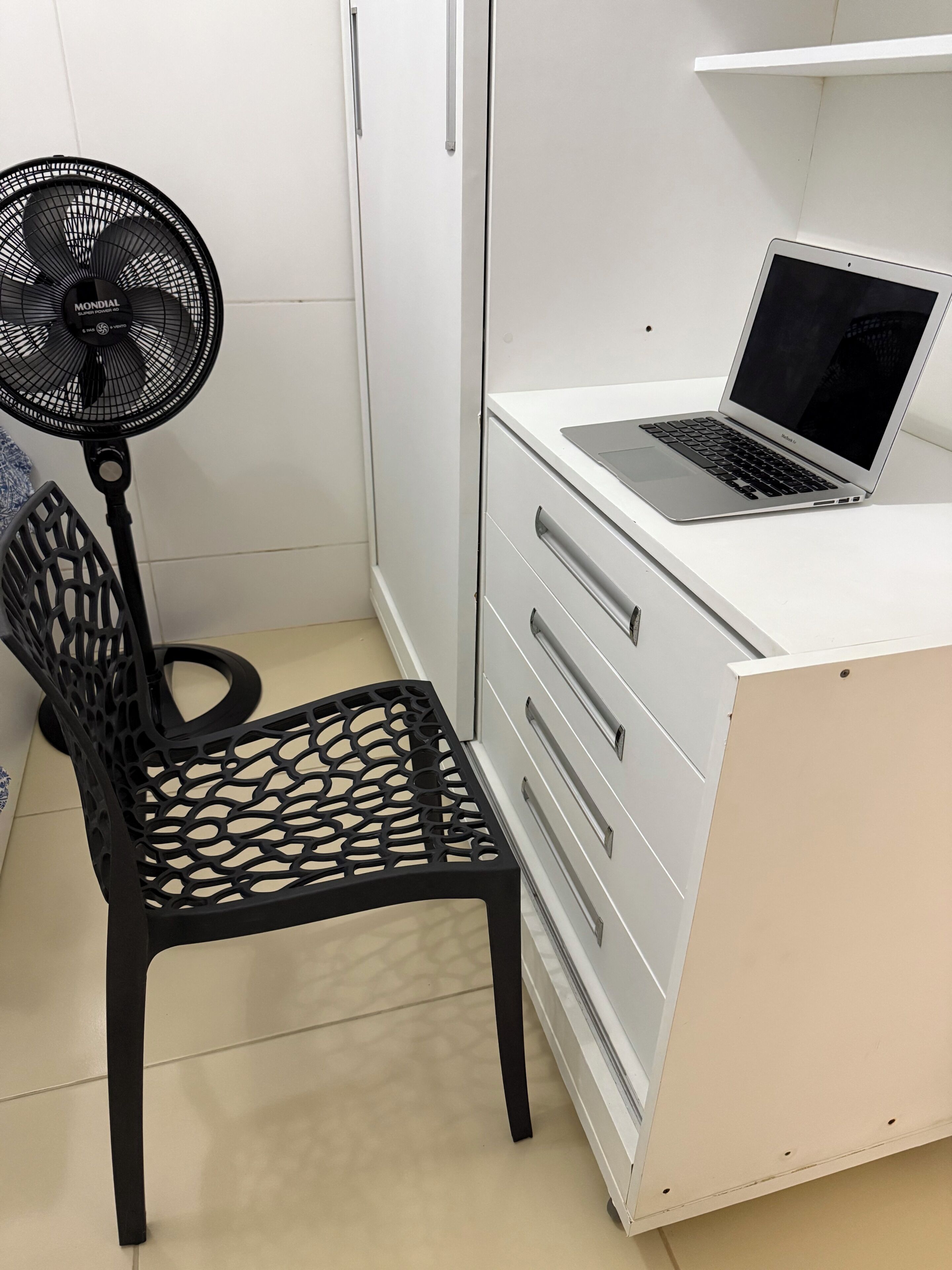 Uma escrivaninha branca com um laptop, uma cadeira preta e um ventilador.