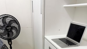 A white desk with a laptop, a black chair, and a standing fan.
