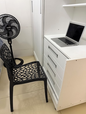 Una taula blanca amb un ordinador portàtil, una cadira negra i un ventilador de peu.