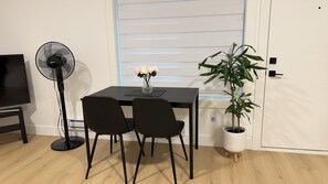 Et værelse med et spisebord, to stole, en ventilator, et fjernsyn, en potteplante og et vindue med persienner.