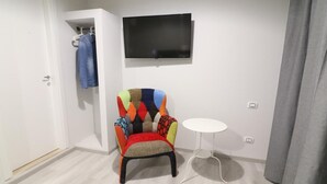 Et værelse med en farverig lænestol, et tv ophængt på væggen, et lille hvidt bord og en dør med en ophængt beklædningsgenstand.