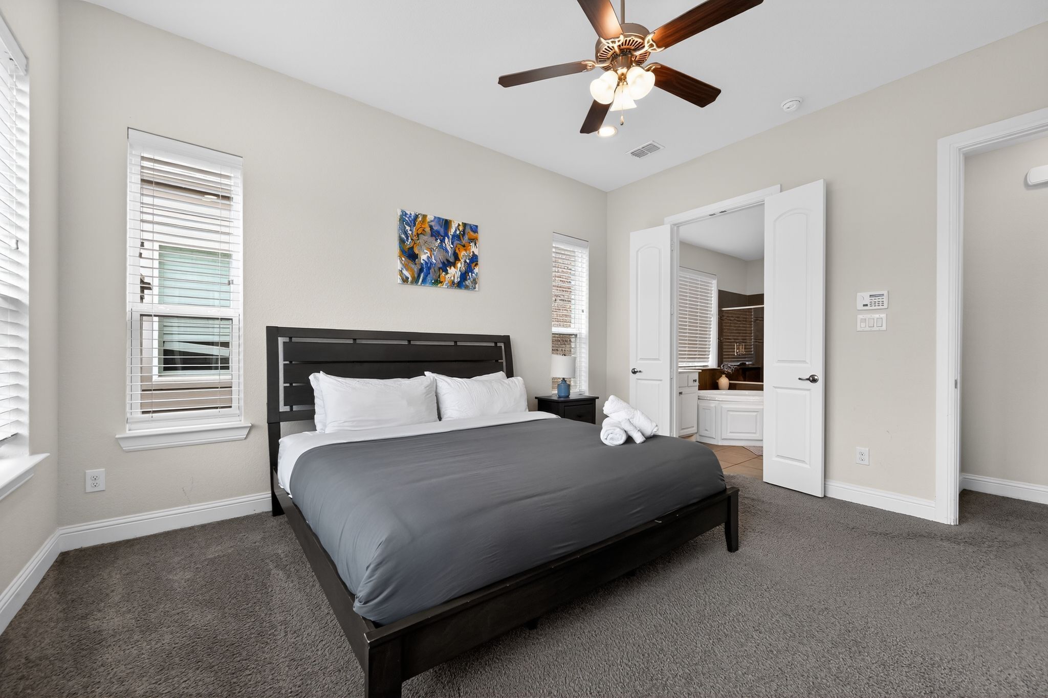 A bedroom with a bed, a ceiling fan, a painting, and a bathroom visible through an open door.