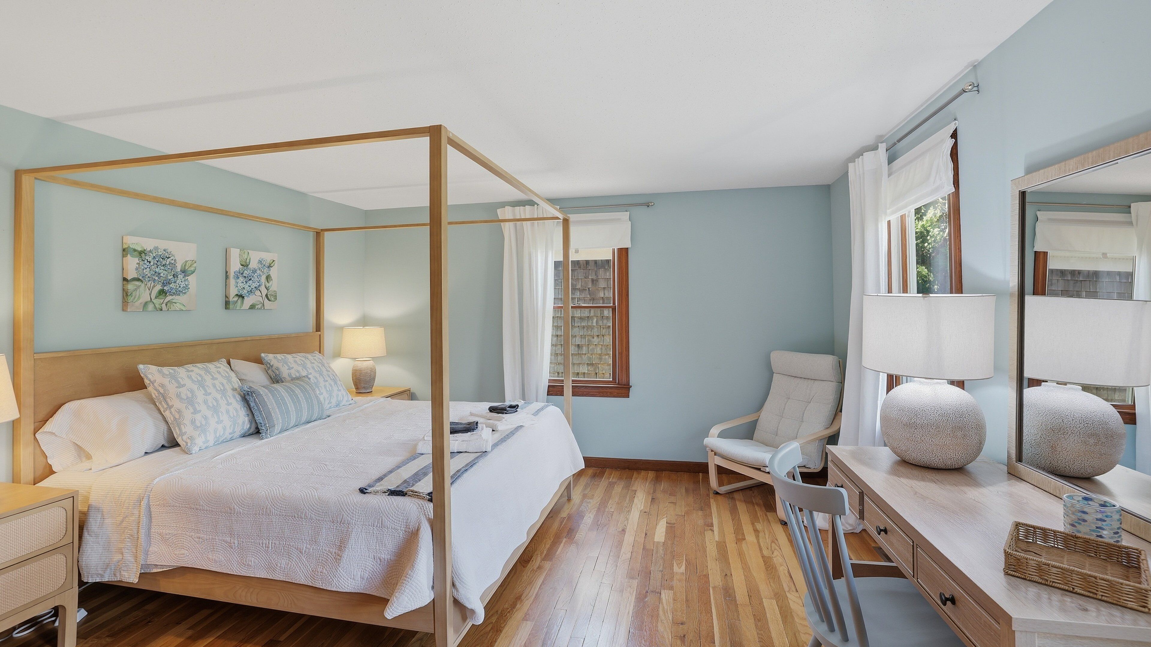 A four-poster bed with a wooden frame, a bedside table with a lamp, a chair, a mirror, and artwork on the wall.