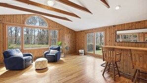 Ein Zimmer mit Holzwänden, großen Fenstern, einer Holzbalkendecke, einer Couch, einem Sessel, einem kleinen Tisch und einer Bar.