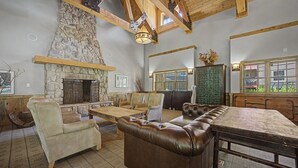 A spacious living room with a stone fireplace, leather sofa, wooden dining table, and a view through a window. - Collins Lake Mountain Condo #24| Pool, Sauna & Spa (Government Camp)