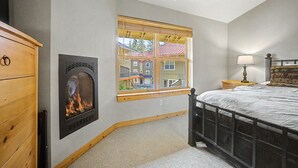 A bedroom with a fireplace, a bed, a nightstand, and a window with a view of buildings. - Collins Lake Mountain Condo #24| Pool, Sauna & Spa (Government Camp)