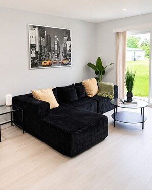 Una sala moderna con un sofá seccional negro, una mesa de centro de vidrio y una pintura de un paisaje urbano en la pared.