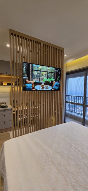A bedroom with a bed, a TV mounted on a wooden panel, a dining area with a table and chairs, and a balcony with a railing. - GRU SKY Studio, 26th floor, New, High Standard, Rooftop, 10 minutes from the Airport (Guarulhos)