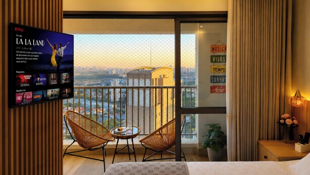 Una habitación de hotel con balcón, un televisor mostrando una película y una cama con colcha estampada.