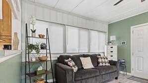 A living room with a gray sofa, a wooden ladder shelf with decorative items, a white door, and a window with blinds.