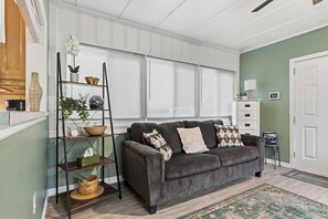 A living room with a gray sofa, a wooden ladder shelf with decorative items, a white door, and a window with blinds.
