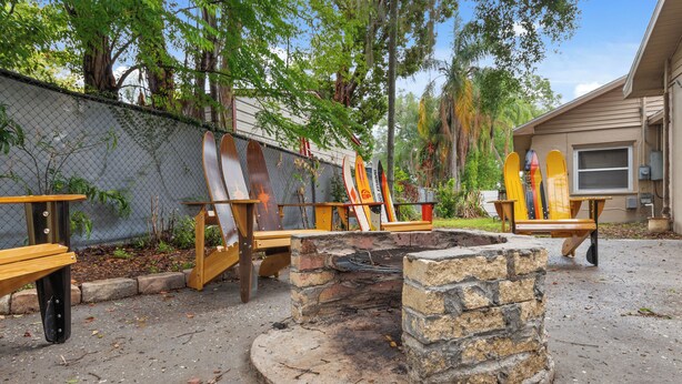 A stone fire pit surrounded by wooden benches, with a chain-link fence and trees in the background. - "Chalet" lake house/walking path/fenced yard/dock (Winter Haven)