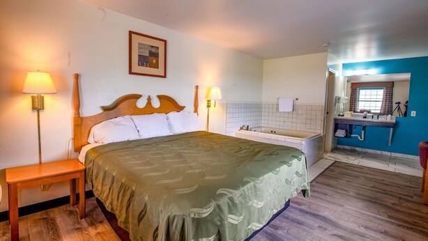 A hotel room with a bed, bedside lamps, a wooden nightstand, a bathroom with a bathtub, and a mirror. - Economy Inn & Suites (Rantoul)
