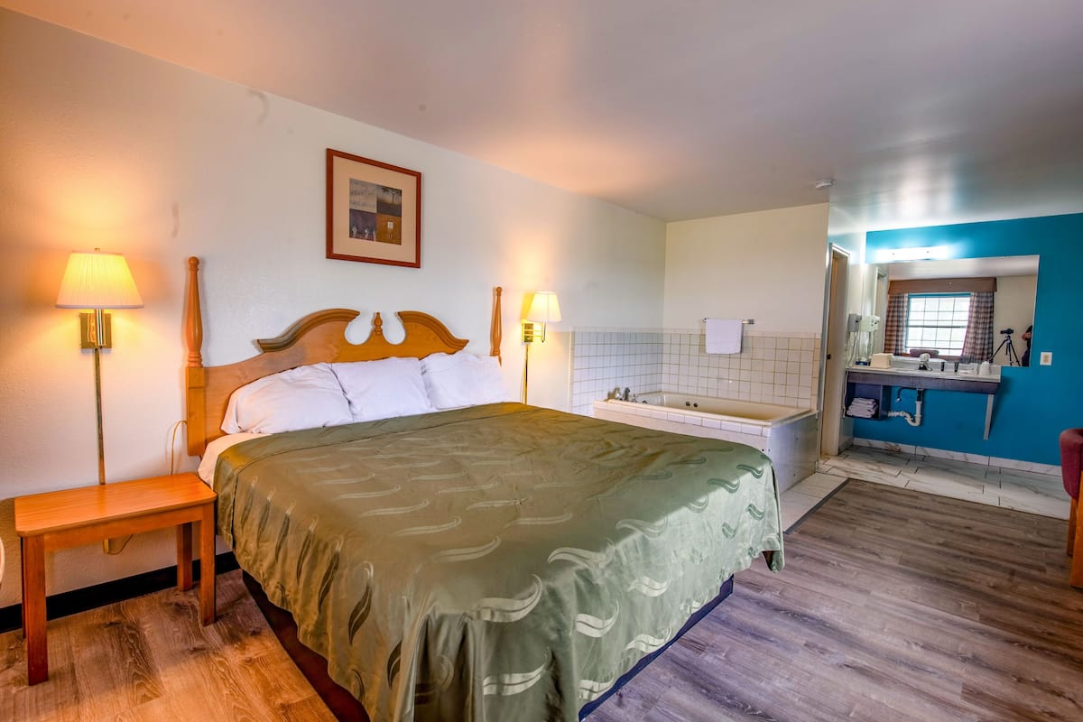 Une chambre d’hôtel comprenant un lit, des lampes de chevet, une table de nuit en bois, une salle de bain avec une baignoire et un miroir.