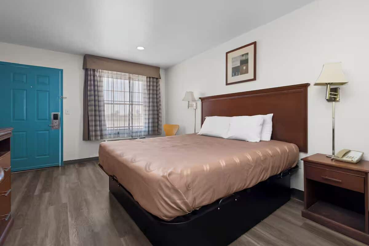 Une chambre d’hôtel comprenant un lit, un bureau, une chaise, une fenêtre avec des rideaux, une porte et une lampe fixée au mur.