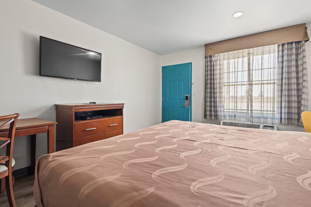Een hotelkamer met een bed, een bureau, een televisie, een blauwe deur en een raam met gordijnen.