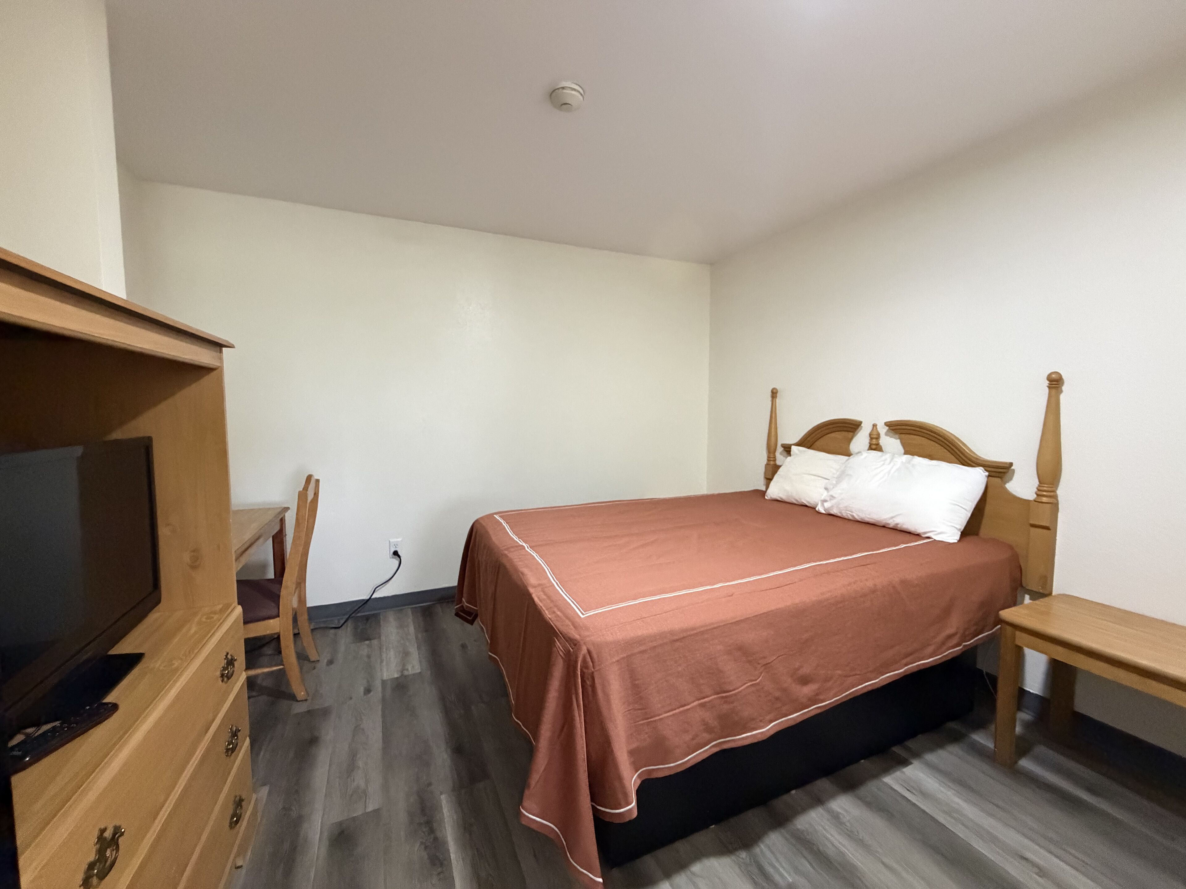 A bedroom with a bed, a wooden dresser, a television, and a desk.