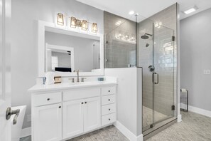 Un baño moderno con ducha envuelta en vidrio, un tocador blanco con cajones y un espejo grande.