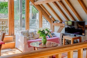 Ein gemütliches Zimmer mit Holzbalkendecke, einer Couch mit Kissen, einem Glastisch mit Vase und einem Fernseher.