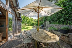 Ein Holztisch mit einem großen Sonnenschirm, umgeben von Korbstühlen, in einem Garten mit Steinmauern und viel Grün.