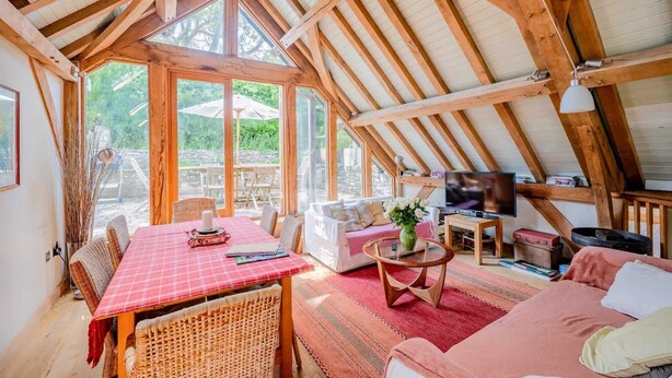 A cozy attic room with a high ceiling, wooden beams, a dining table, a sofa, a TV, and a view of greenery outside. - Airy Barn Conversion, Private Parking & Garden (Burrington)