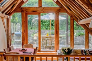 Ein gemütliches Zimmer mit Holzbalkendecke, großen Fenstern, einem Esstisch und Korbmöbeln.