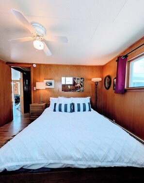 A bedroom with a bed, a ceiling fan, wooden walls, a window with a purple curtain, and a hallway leading to another room. - Cozy / River Front / Charm (Salida)