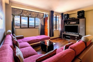A living room with a sofa set, a flat-screen TV, speakers, and a view of a mountainous landscape through the windows. - Incredible views of the Andes | Central location & Laundry & Private parking (Cusco)