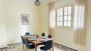 A dining room with a wooden table set for six, six chairs, a potted plant, and a window with curtains. - Villa plage David Bouznika (Bouznika)
