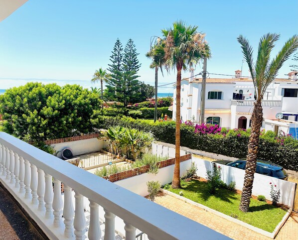 A residential area with white buildings, a balcony, and a garden with palm trees and shrubs.