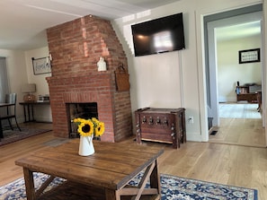 Ein Wohnzimmer mit Kamin aus Ziegelsteinen, einem Holztisch mit Sonnenblumen in einer Vase, einem Flachbildfernseher und einem Schild mit der Aufschrift 'Welcome to the Mountain Brewers'.