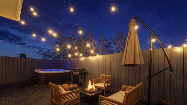 Een terras buiten met een hottub, barbecue en zitgedeelte, verlicht met lichtslingers.