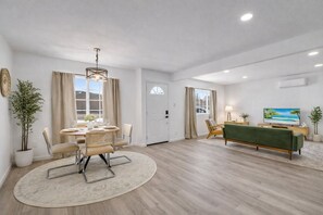 A modern living room with a dining area, a green sofa, a TV, and a large window. - Joshua Tree Oasis - 10 Min to Park - Hot Tub, Fire Pit & Stargazing (Joshua Tree)